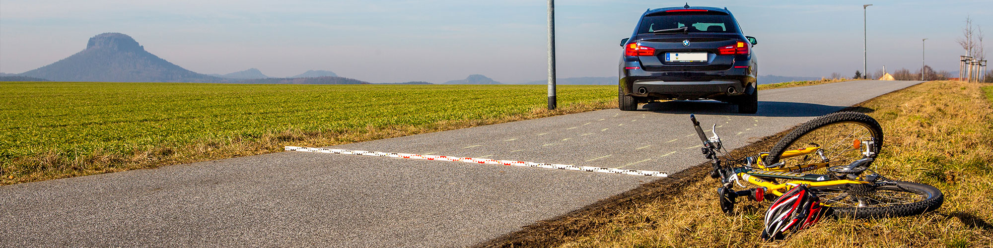 Im Vordergrund liegt ein Fahrrad neben der Fahrbahn auf der Straße, ein Stück weiter auf der Straße steht leicht schräg ein Auto, auf der Straße liegt eine Messlatte und die Spuren des Autos wurden mit Kreide markiert